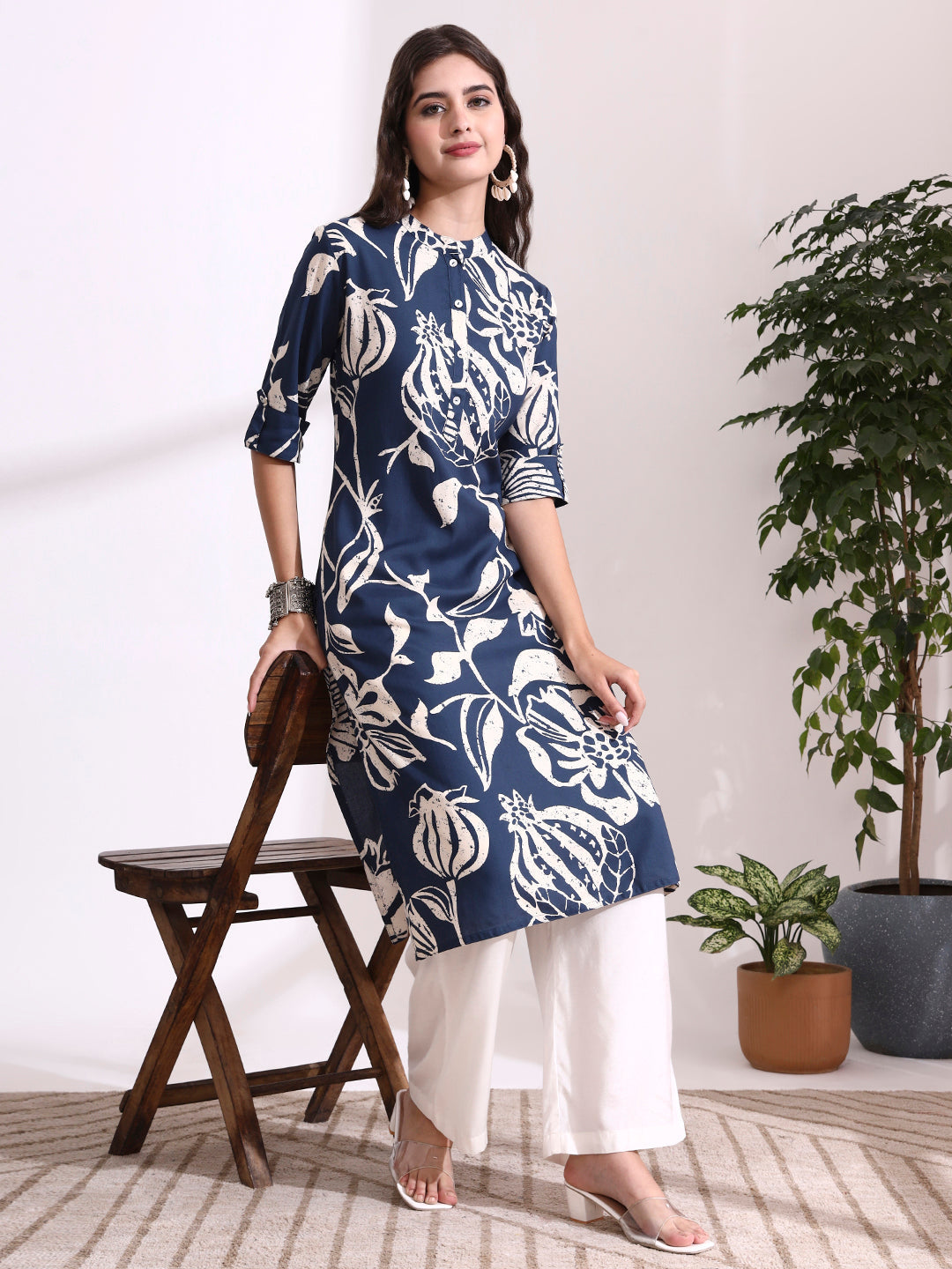 Woman wearing a blue and white patterned kurta with white pants, standing in a room with plants.