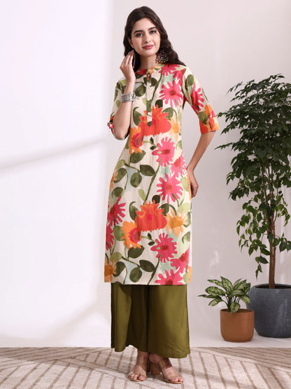 Woman wearing a floral kurta with green pants standing indoors next to a plant.
