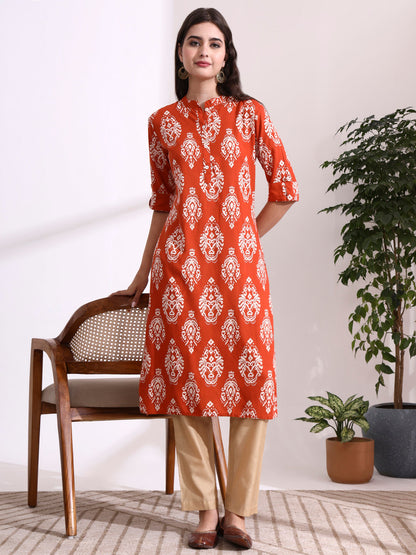 Woman wearing a red and white patterned kurta with beige pants in a room with a chair and plants.